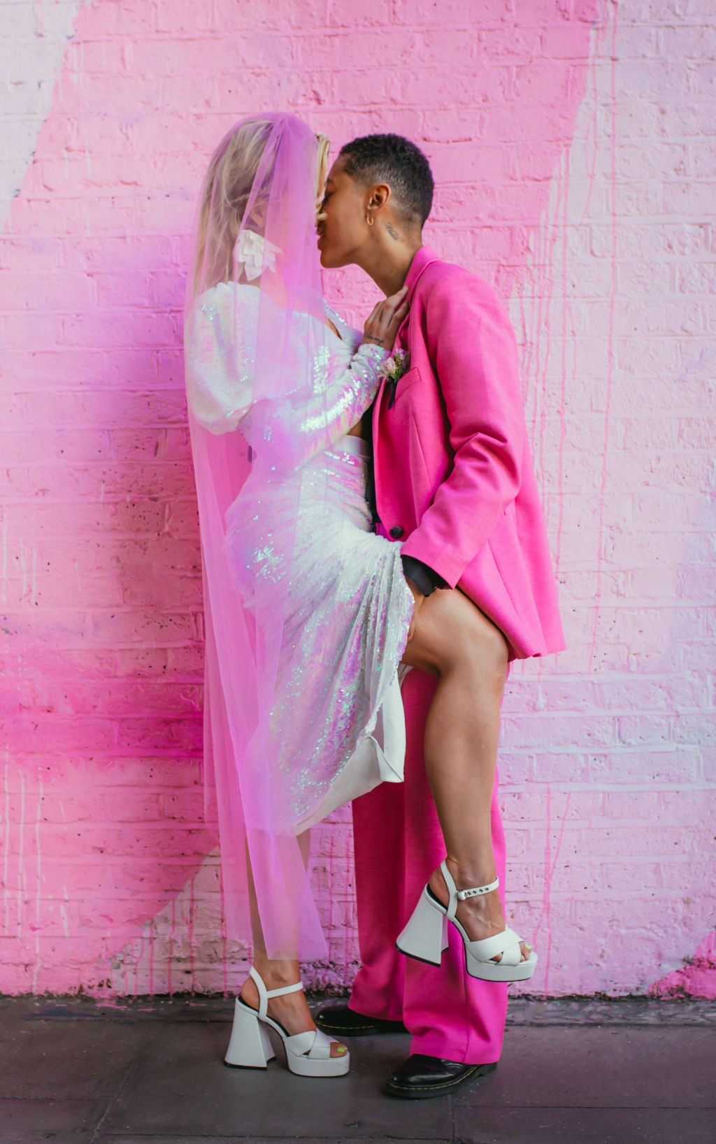 a bride with statement earrings, an iridescent, sequinned 2 piece wedding dress and loose tousled hair and a groom with a grade 1 hair style wearing a bright pink suit, black shire and gold jewellery 