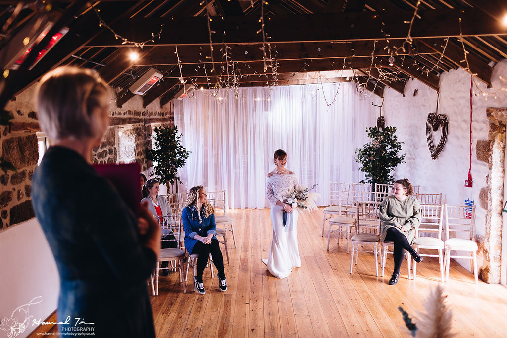 Celebrant view of the bride walking down the aisle at Chypraze Barn holding a large dried floral bouquet surrounded by white wash chiavari chairs