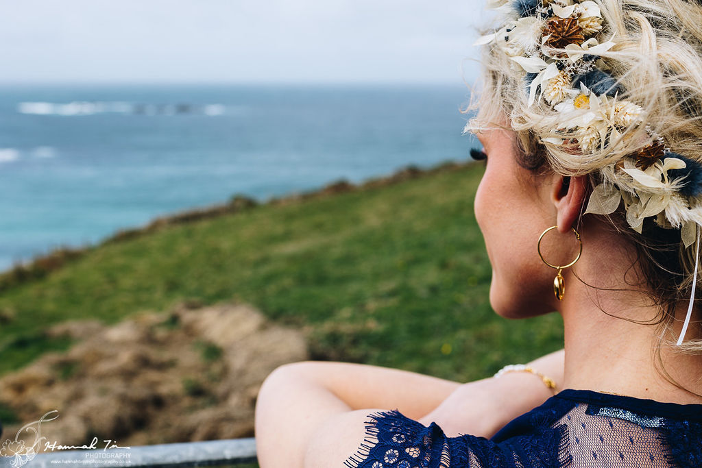 Image of bridesmaid wearing a royal blue bridesmaid dress looking out at the Cornish Coastline. with hanging gold earrings and a dried flower floral crown